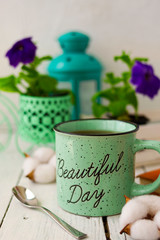 Bright still life with tea, cotton bolls, a book, a spoon and flowers with a lantern on the background. On the mug there is an inscription Beautiful day. 