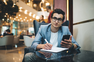 Serious caucasian male executive manager in trendy formal wear and glasses using mobile phone for research while planning meeting in cafe, confident businessman noting information about banking