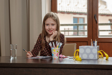 Close up of cute blond hair school girl, sitting at the desk with school supplies, drawing, looking at the camera, homeschooling, closed at home during quarantine 2020
