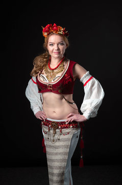 beautiful woman in traditional slavic dance costume with golden necklace, circlet of flowers, red west, white sleeves and skirt looking at camera and holding hands on hips isolated on black