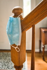 Mask hanging on stair railing looking towards entrance of home. Symbolic of need to wear face mask when leaving home for protecting from COVID-19