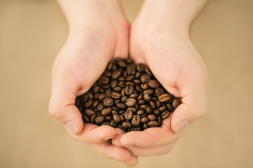 A person is holding a large handful of quality aromatic roasted coffee beans on a beige background, illuminated by light.