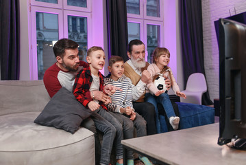 Agitated attractive three generations of people as dad,grandpa and grandchildren which all sitting on the couch at home and enjoying at soccer match and shouting when see scored ball