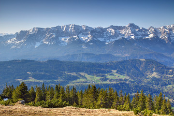 Steep snowy Wettersteinwand rock face and Dreitorspitze peak above woods, glades and Wamberg village in sunny morning from Wank summit, Wetterstein range Northern Limestone Alps Bavaria Germany Europe