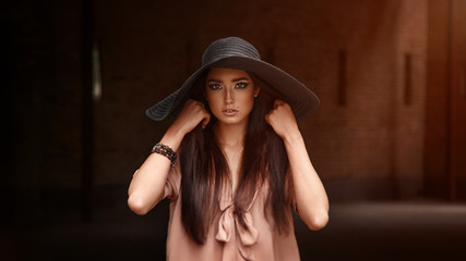 Outdoor fashion portrait of young fashionable brunette woman, model wearing black hat, posing at sunset.