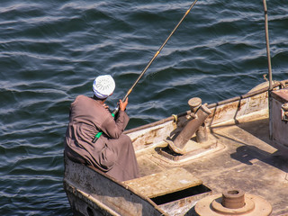 Man fishing in an old commercial fishing boat wearing a white hat and brown robe, in Luxor, Egypt.