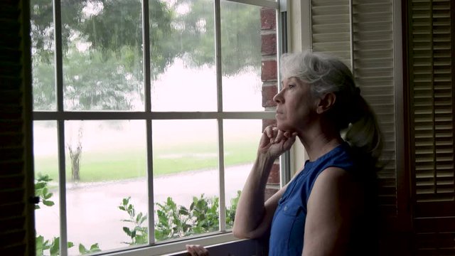 An older melancholy woman stands looking out of a window at a torrential rainstorm.
