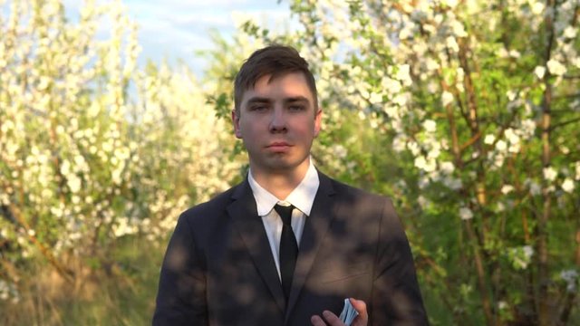 A businessman is standing in the garden and waving bundles of dollars, and then points his hands to the garden behind him. It became hot men in a suit in nature.