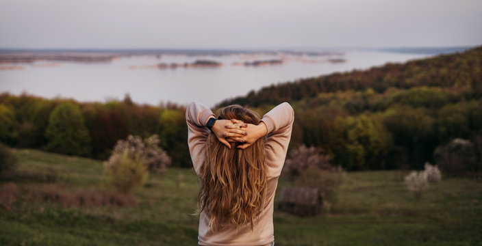  A young girl stands with her back with blond hair and hands behind her head. Looks at the horizon and the river behind the hill at sunset. Panorama. Loneliness. A look into the future. Hope