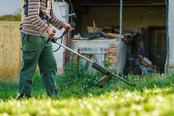 Young caucasian man farmer gardener standing in the field with string trimmer petrol Brushcutter cut weed grass working on farm cutting in the field in sunny day by the house in the village side view