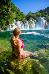 Blonde girl with long attached hair wearing a pink bikini bathing in the transparent waters of Krka National Park under the famous waterfall in a lush environment