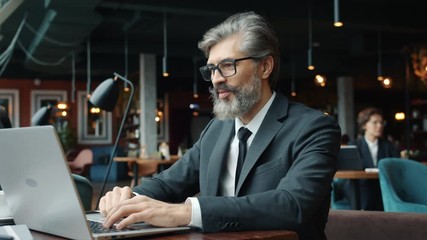 Mature businessman is making online video call talking and gesturing using laptop in cafe. Modern gadgets, technology and communication concept.