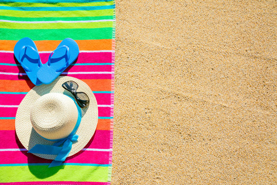 Beach towel with hat, sunglasses and flip flops photographed from above on sandy beach, hot summer day accessories, vacation destination, sunny tropical beach background