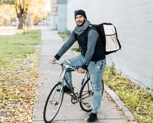 A young male with blue eyes who works for a take away food company poses with his bike and food bag.