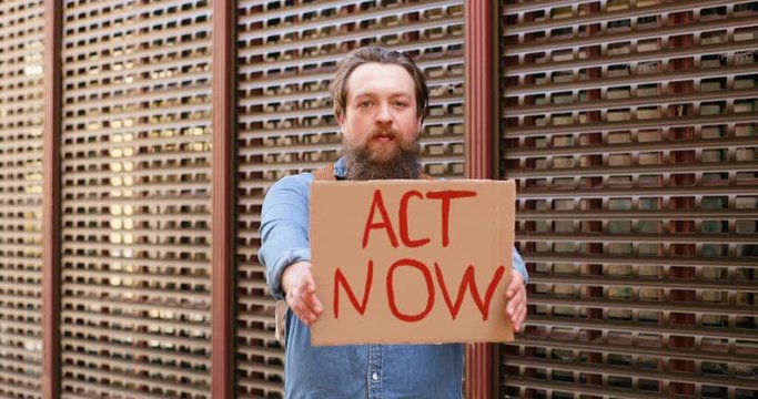 Portrait shot of Caucasian young man demonstrating poster with words Act Now. Male activist showing board with protest on political or environmental issues. Lonely protesting. Activism concept.