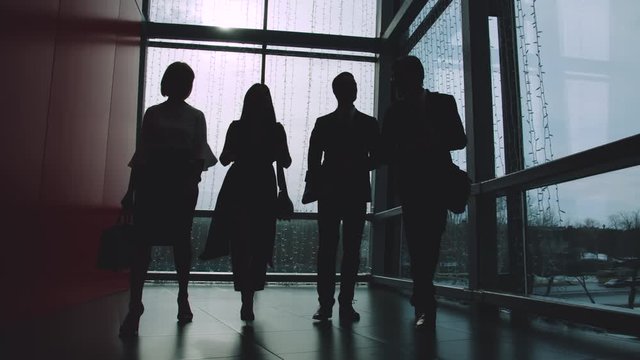 Silhouettes of men and women walking in office hallway against window and talking discussing work and business. Businesspeople and workspace concept.