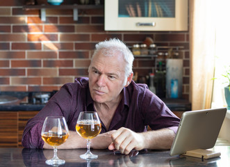 An irritated man with a gadget and two glasses of wine is sitting in the kitchen