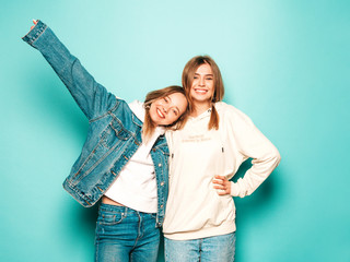 Two young beautiful brunette smiling hipster girls in trendy summer hoodie and jeans jacket clothes. Sexy carefree women posing near blue wall. Trendy and positive models having fun