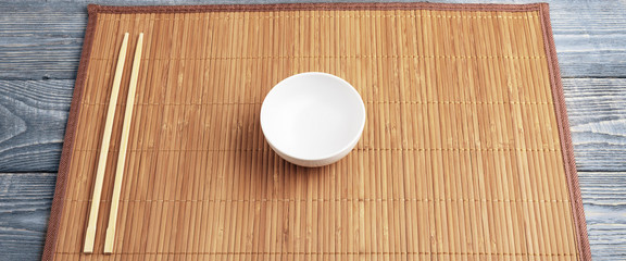Two wooden chopsticks and a white cup on a bamboo mat