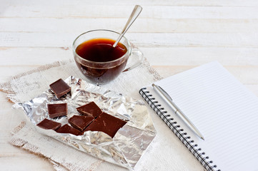 A cup of coffee, chocolate, a sheet of paper and a pen on a white wooden surface. Background for...