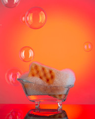 Sponge for washing dishes and cleaning in the foam and in a tiny bath suurrounded of soap bubbles on red background with reflection.