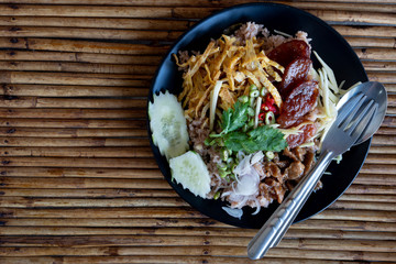 Thai food Rice Seasoned with Shrimp Paste or Khaao Khlook Gabpi. Put on a black plate and have a spoon and fork. With rattan stalks arranged in the background.