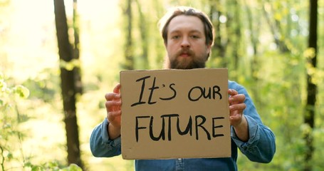 Portrait shot of Caucasian young male eco acivist standing in green forest and holding poster with words It's our Future. Handsome man protesting for clean and safe environment.