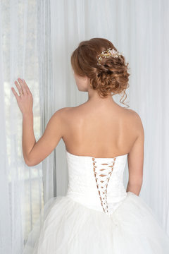 A bride with a beautiful haircut with decoration wearing  white wedding dress looks dreamily out the window