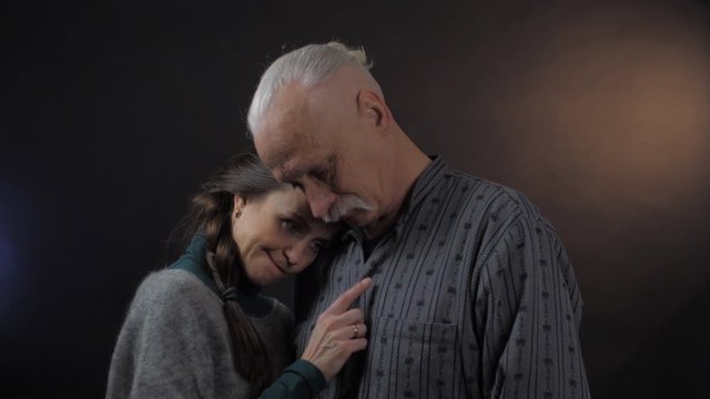 lovely dark haired lady holds head on elderly man shoulder speaks modestly and smiles against dark background close view. Concept people emotion