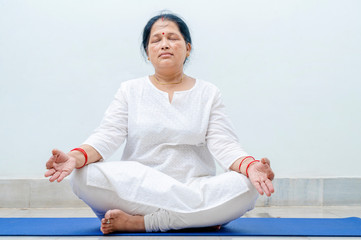 Middle Aged or an Old Indian Woman Performing Yoga Early Morning, in her house in white dress. Stay Home Stay Safe and Fit