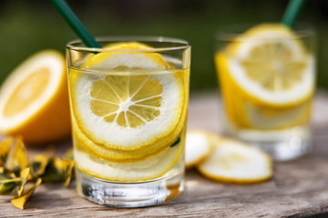 Summer drink, glasses of water filled with slices of lemons, stand on a wooden board on the lawn in sunny warm day, the background is blurred, shallow depth of field, selective focus.