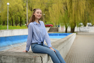 Fototapeta na wymiar Photo of a young pretty girl with a smile in a blue shirt and jeans. Model posing sitting in spring park outdoors in the city.