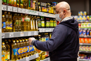 A man in a medical mask and gloves with a bottle of vegetable oil in a supermarket. Big choice. Precautions during the coronavirus pandemic.