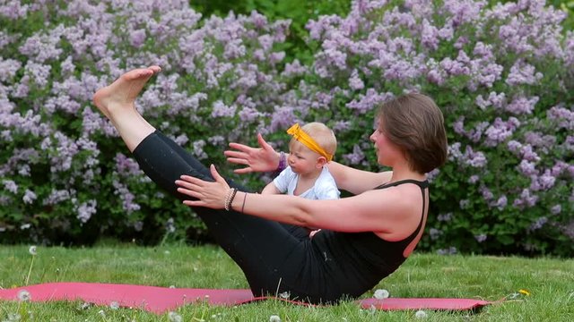  CZ, Prague, Karlinske nam, 9.3.2020 Outdoor yoga with baby. Young woman, does gimnastic exercises in park near lilacs plant. Mother with daughter. Natural parenting lifestyle.