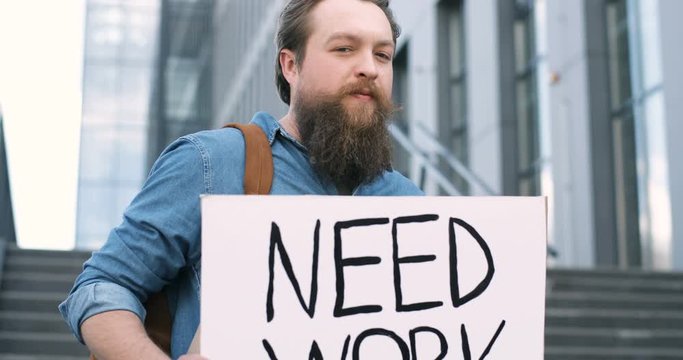 Close up of Caucasian young jobless man with beard standing outdoor at big stairs and showing table with words Need Work. Male workless demonstrating board with protest against unemployment.