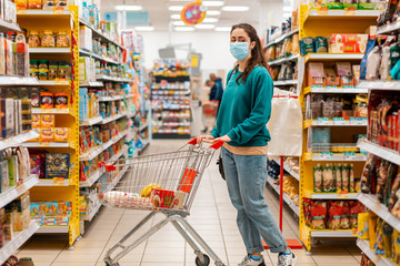 Shopping. A young Caucasian woman with a medical mask on her face, rolls a grocery cart through the aisles of the store. Side view. The concept of buying products and the coronovirus pandemic