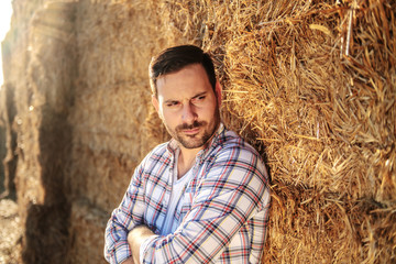Naklejka na ściany i meble Handsome caucasian serious farmer standing outdoors with arms crossed and leaning on bales of hay.