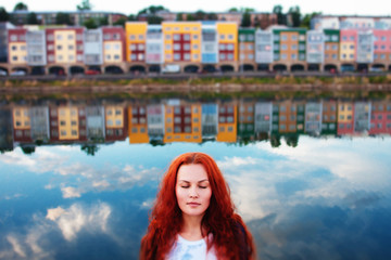 Fototapeta na wymiar Portrait of an attractive red-haired girl on the background of a river with a reflection of colored houses