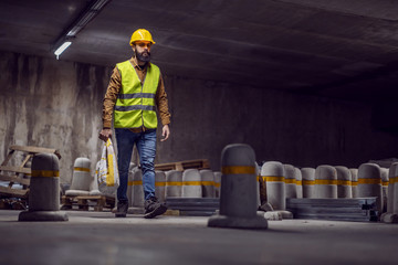 Full length of attractive hardworking bearded worker carrying sack with concrete while walking in...