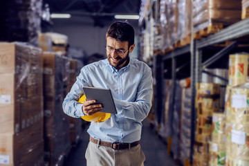 Young handsome bearded employee holding protective helmet under armpit and using tablet to check out goods.