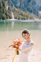 Beautiful bride in a white dress with sleeves and lace, with a yellow autumn bouquet of dried flowers and peony roses, on the Lago di Braies in Italy. Destination wedding in Europe, on Braies lake.
