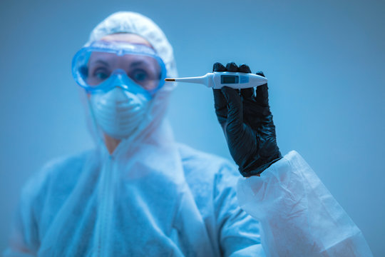 Medical Doctor, Scientist In Full Body Suit For Prevention From Viruses And Diseases Holding A Thermometer For Body Temperature.