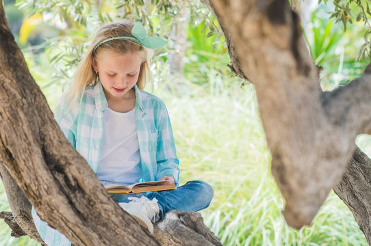 Blonde girl reading a book on a large spreading tree.