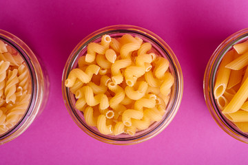 Three glass jars with variety of uncooked golden wheat pasta on minimal pink background, top view macro