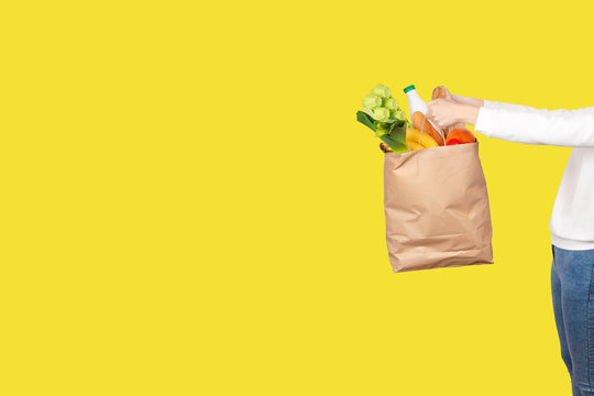 Girl holds a paper bag filled with groceries such as fruits, vegetables, milk, yogurt, eggs isolated on yellow.