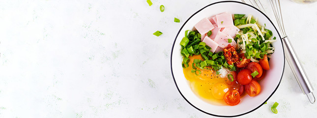 Omelette ingredients: eggs, ham, tomatoes, green herbs, milk and cheese on the light table. Top view, overhead, banner