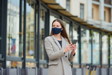 A girl with long hair in a navy blue face mask to avoid the spread coronavirus uses a smartphone in the street. A woman in a face mask against COVID-19 wears a coat looks away in the city.