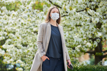 A girl in a medical face mask to avoid the spread coronavirus holds a smartphone in the park. A...