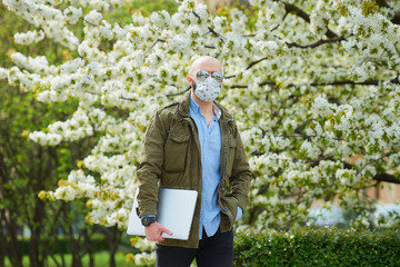 A bald man with a beard in a medical face mask to avoid the spread coronavirus walks with a laptop in the park. A guy wears n95 face mask and a pilot sunglasses in the garden between flowering trees.