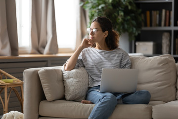 Dreamy young Caucasian girl in glasses sit on couch in living room hold laptop look in window distance dreaming, thoughtful female distracted from computer work dreaming or thinking at home